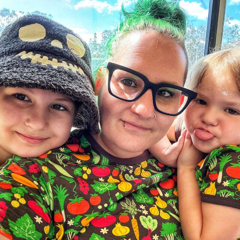 Woman with green hair and two children wearing matching vegetable-patterned clothing outdoors.