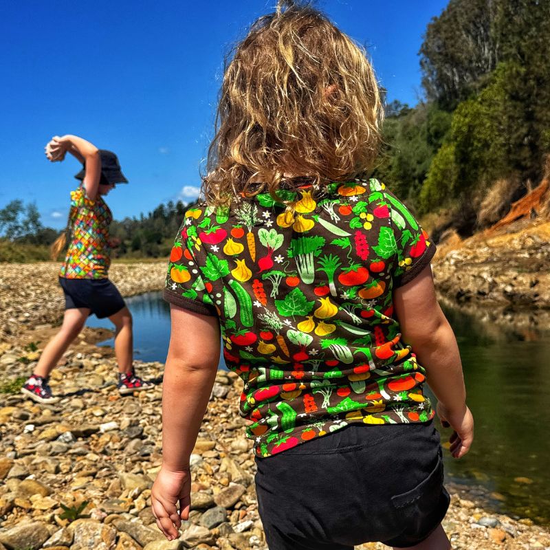 Two children wearing colorful DUNS Sweden t-shirts with vegetable patterns by a river.