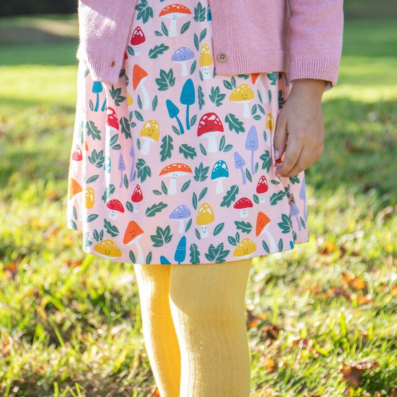 Child wearing a colorful dress with mushroom pattern and pink cardigan in a grassy outdoor setting