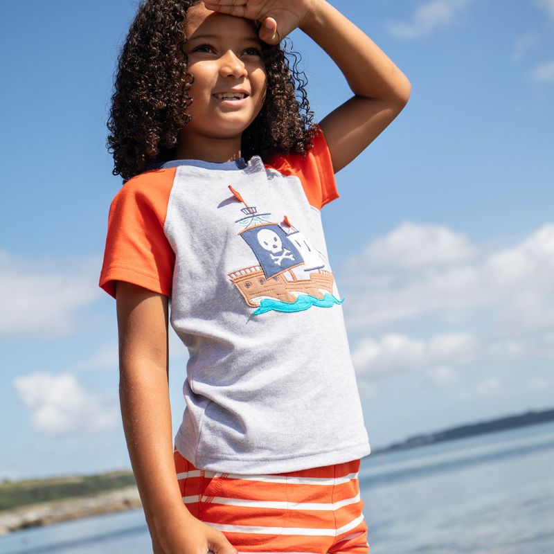 Child wearing a pirate-themed t-shirt with an orange and gray design, standing by a body of water.