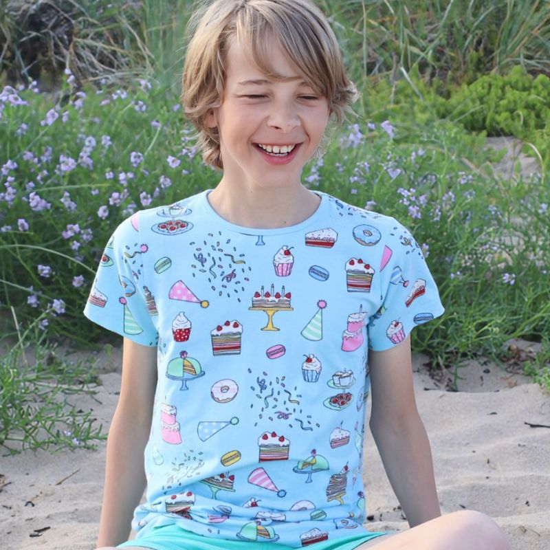 Child wearing a light blue t-shirt with colorful cake designs, sitting on sand with plants in the background.