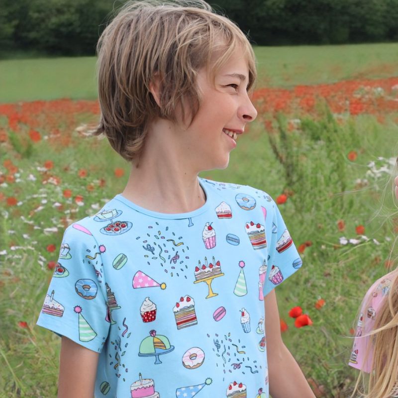 Child wearing a light blue t-shirt with colorful cake designs, sitting on sand with plants in the background.