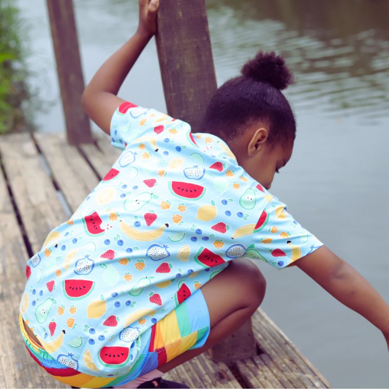 Child wearing a colorful shirt with fruit patterns sitting on a wooden dock by water.