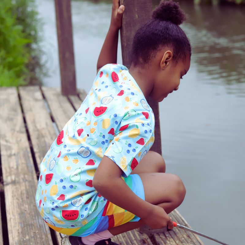 Child in a colorful outfit sitting on a wooden dock by a body of water.