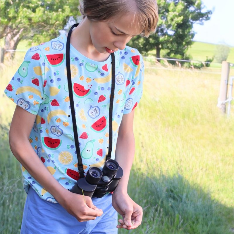 Child holding binoculars and wearing a fruit print Villervalla t-shirt  in a grassy outdoor setting