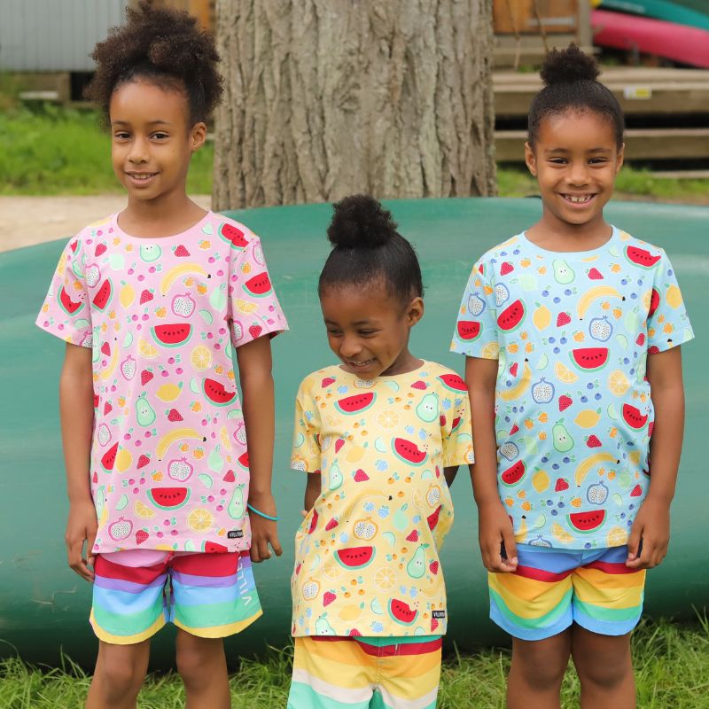 Three children wearing colorful outfits with fruit patterns standing outdoors.