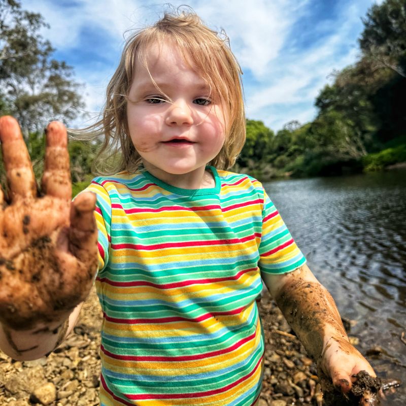 Child in a Villervalla striped shirt standing by a body of water, showing dirty hands.