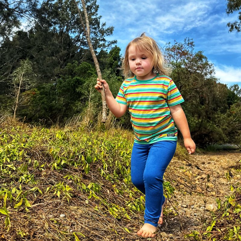 Child in a Villervalla striped shirt and blue pants standing outdoors with trees and grass in the background