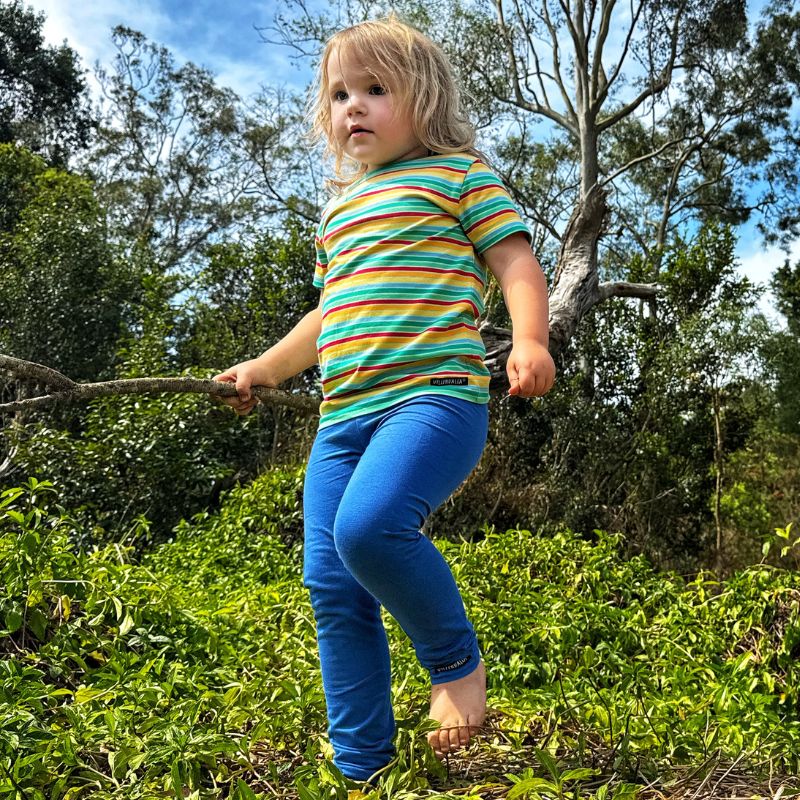 Child in a Villervalla striped shirt and blue pants standing outdoors with trees in the background