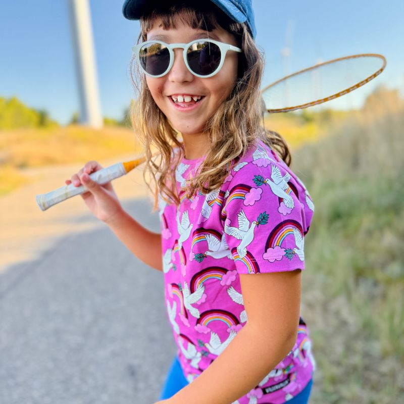 Child wearing a colorful Villervalla Peace t-shirt with rainbow and dove patterns, holding a badminton racket outdoors.