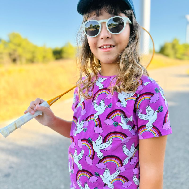Child wearing a colorful Villervalla Peace t-shirt with rainbow and dove patterns, holding a badminton racket outdoors.