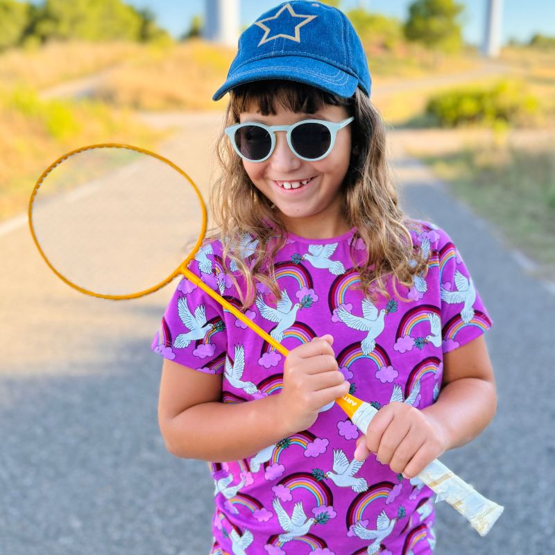 Child holding a yellow badminton racket and wearing a Villervalla Peace t-shirt outdoors on a sunny day